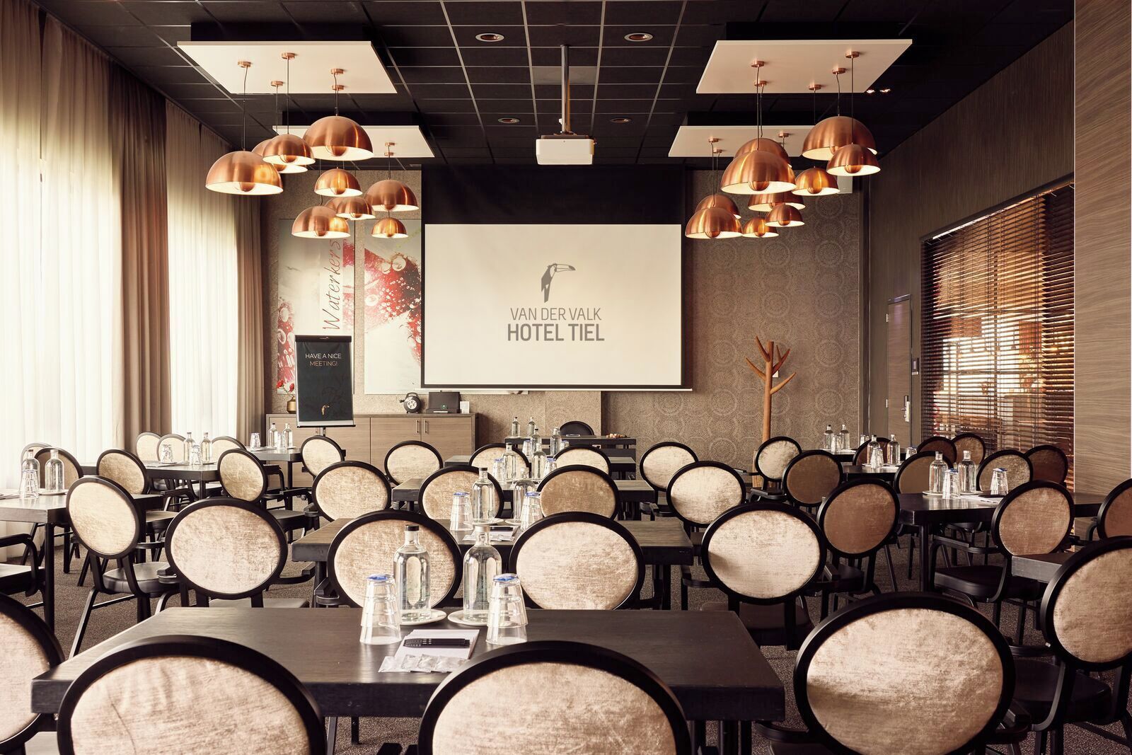 A conference room with multiple tables, chairs, and a large screen displaying 'VAN DER VALK HOTEL TIEL'.