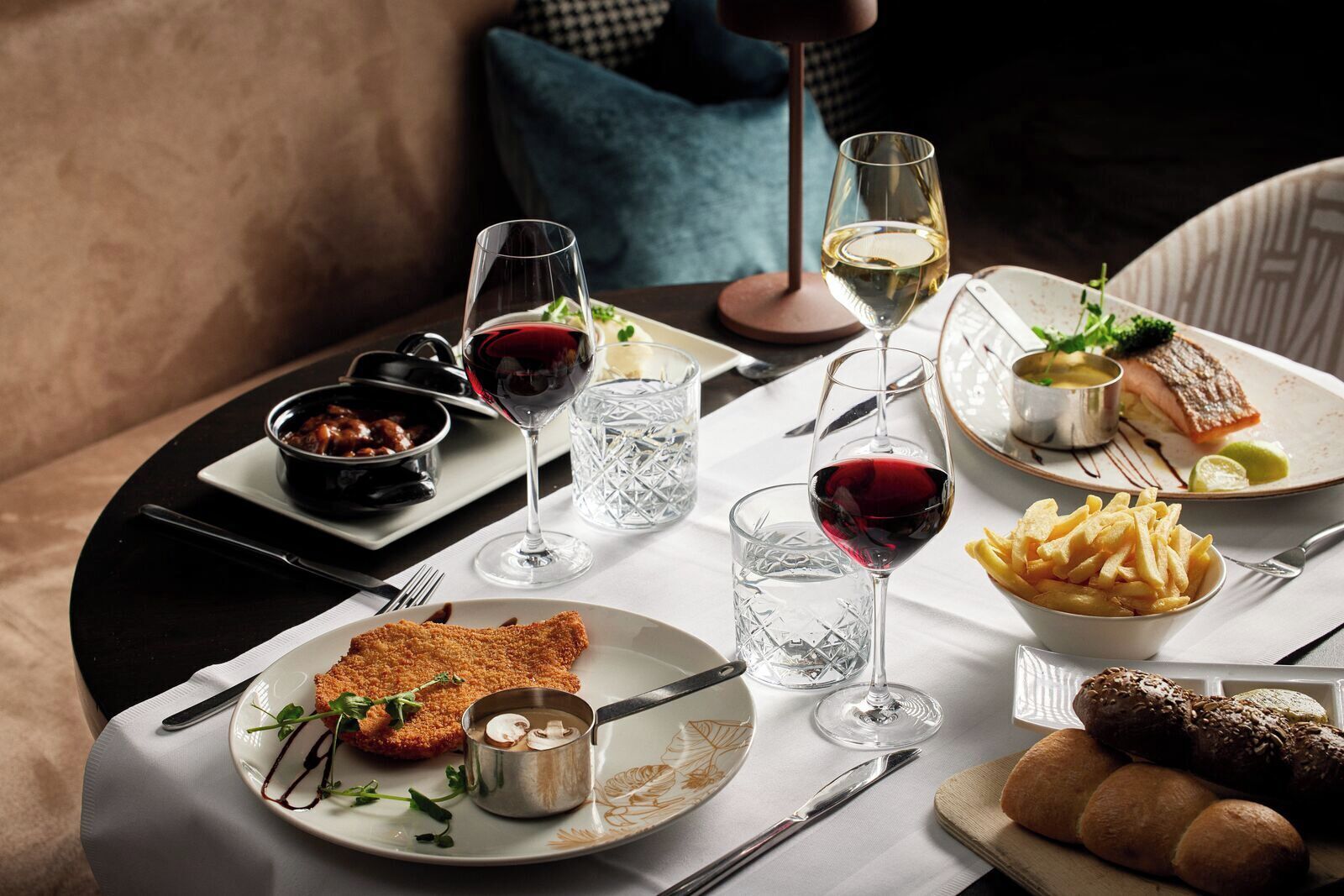 Een eettafel gedekt met diverse gerechten, waaronder een gepaneerde schnitzel, frieten en een visgerecht, vergezeld van glazen rode wijn en water.