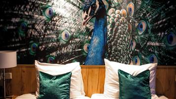 Un dormitorio con una cama, dos almohadas, una mesita de noche, una lámpara y un mural con motivos de pavo real en la pared.