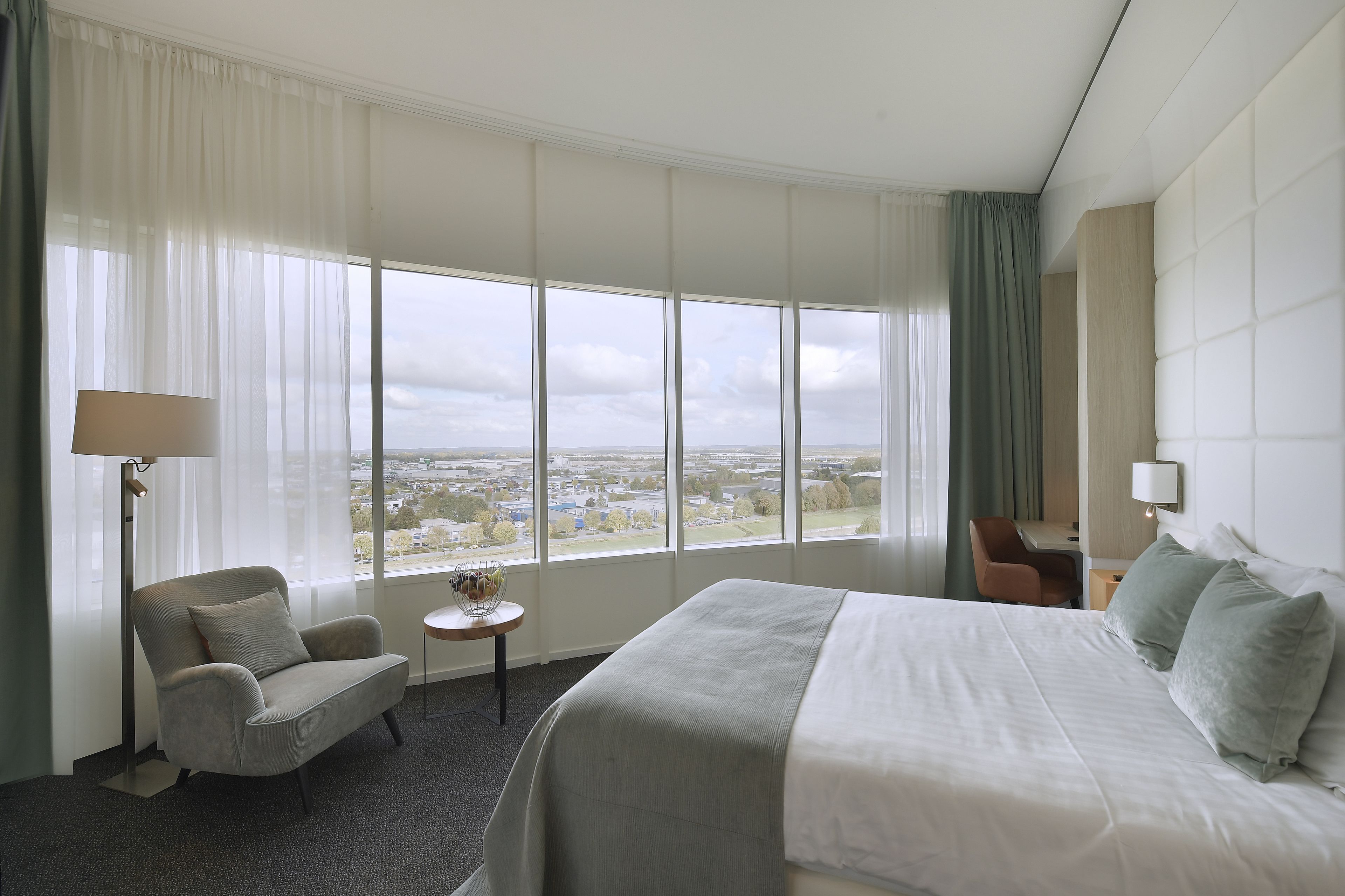 A hotel room with a large bed, a chair, a small table, and a view of the city through large windows.