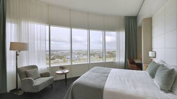 A hotel room with a large bed, a chair, a small table, and a view of the city through large windows.