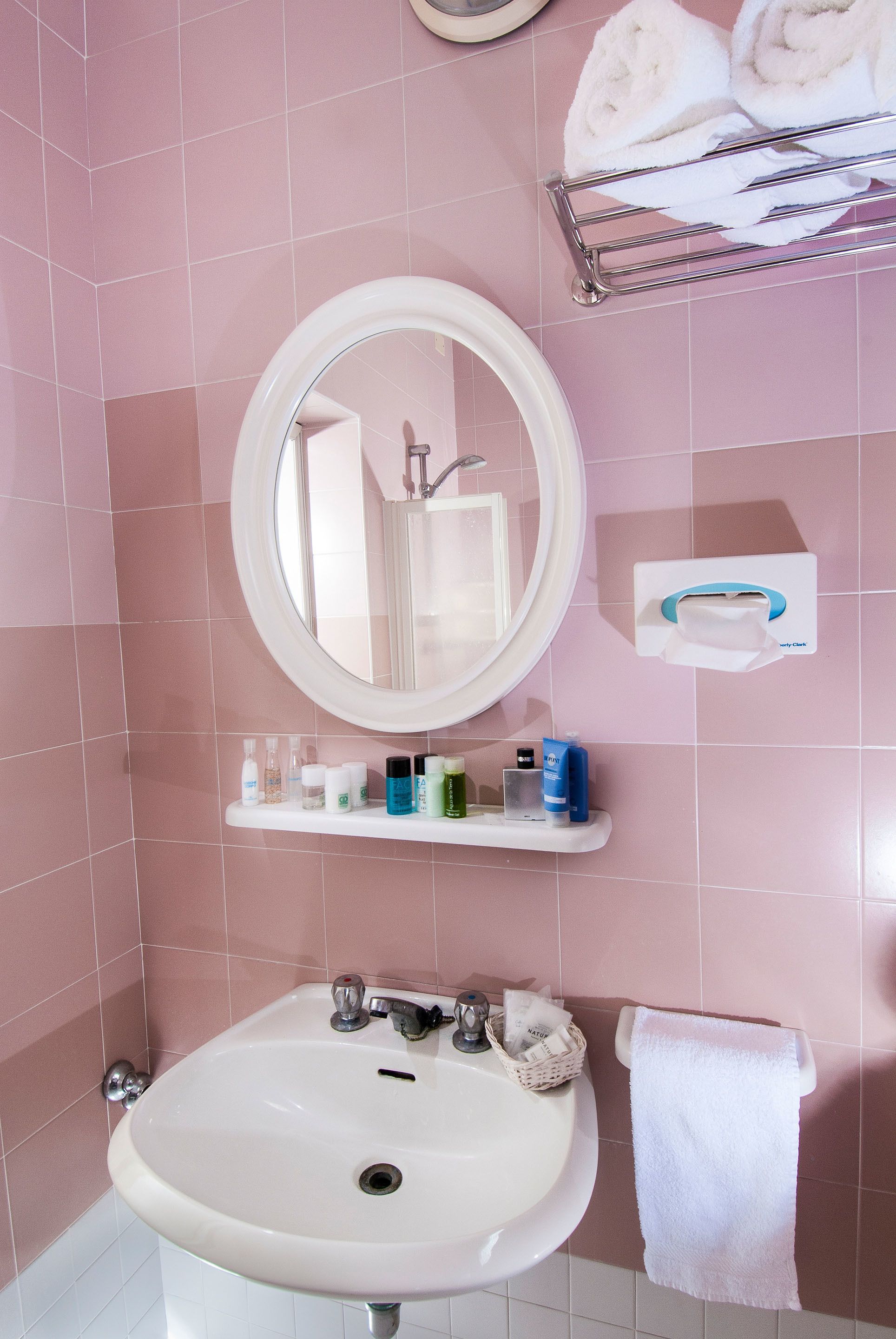 Un baño con azulejos rosas, un lavabo blanco, un espejo redondo y un perchero con toallas enrolladas.
