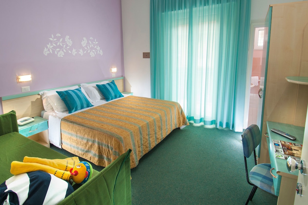A hotel room with a bed, a desk with a chair, a green sofa, a plush toy, and a window with curtains.