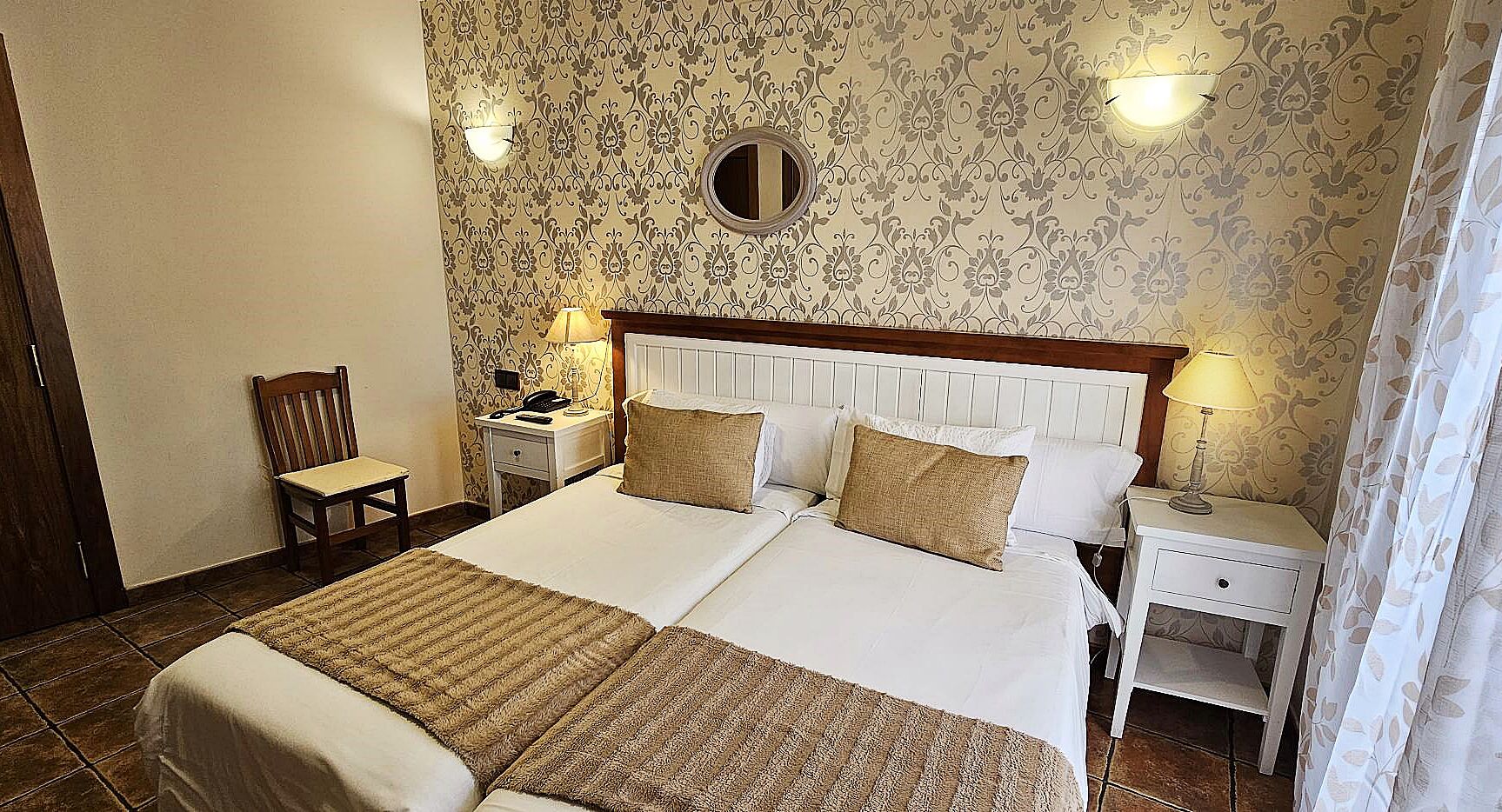 A double bed with two pillows, a bedside table with a lamp, a chair, and a mirror on the wall.