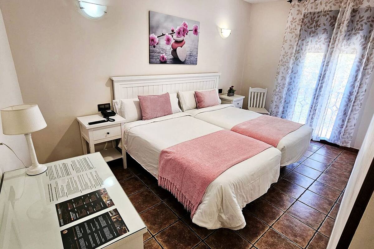 A hotel room with two beds, a desk with a lamp, a nightstand, a wall-mounted phone, and a painting above the bed.