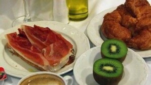 Una mesa de desayuno con jamón serrano, rodajas de kiwi, croissants, un vaso de zumo de naranja y una taza de café.