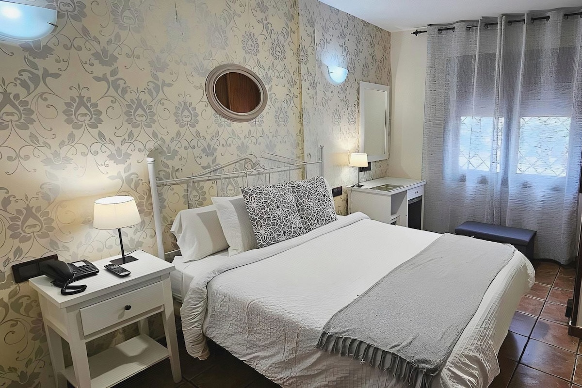 Quarto de hotel com cama, mesas de cabeceira, escrivaninha, espelho, janela com cortinas e parede com padrão floral.