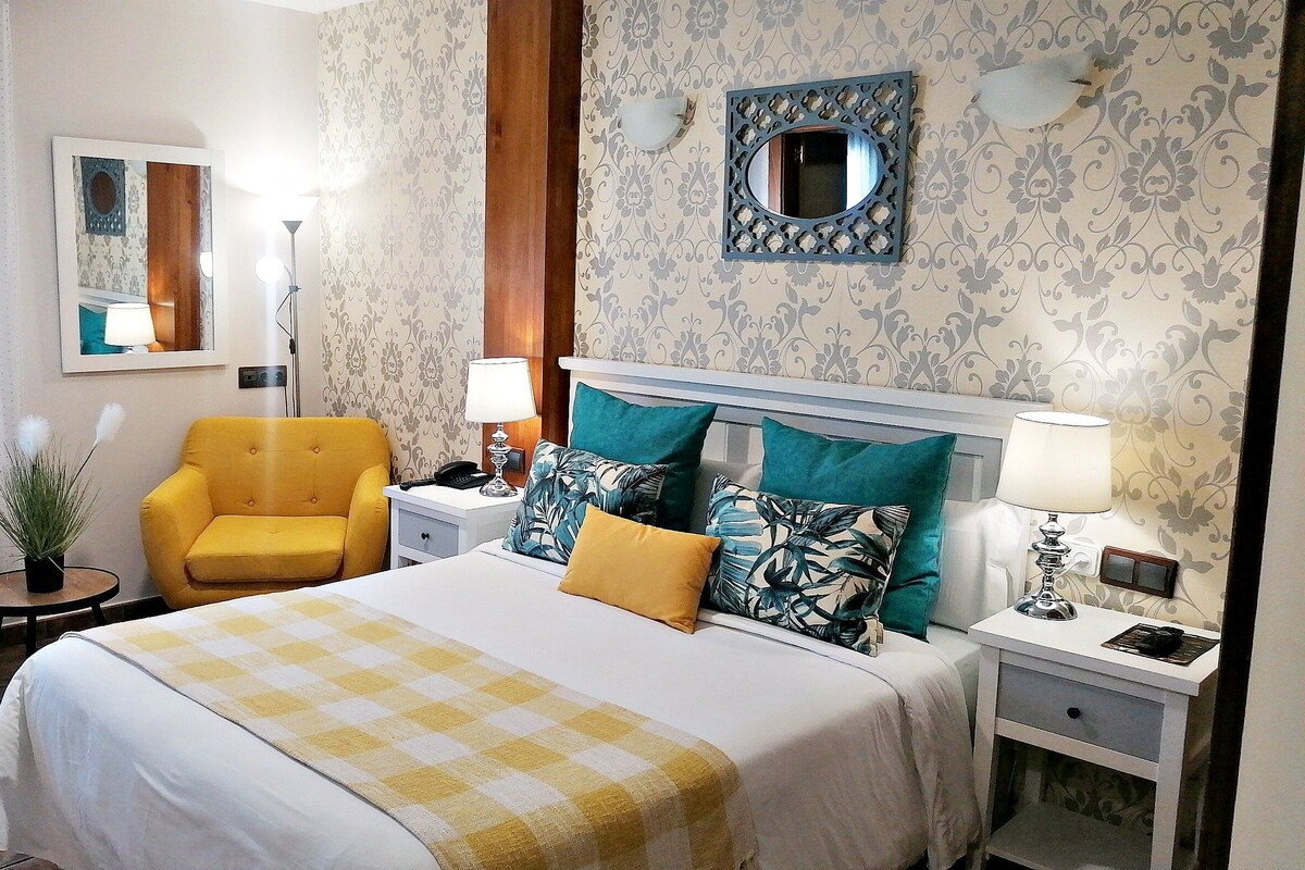 Quarto de hotel com cama, uma cadeira, mesa de cabeceira, abajur, espelho e parede com papel de parede estampado.