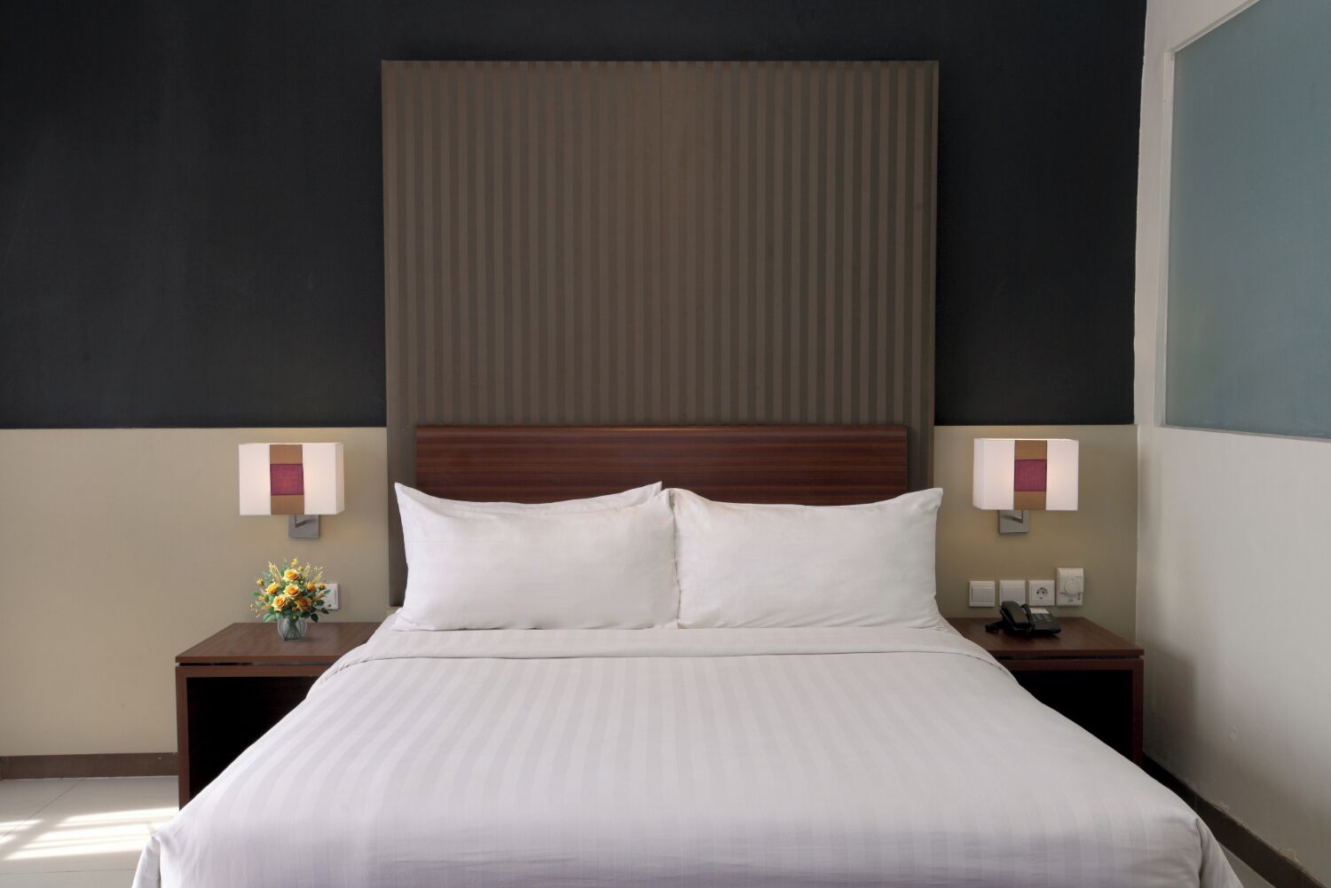 A hotel room with a large bed, two bedside tables, a nightstand, and a wall-mounted lamp.