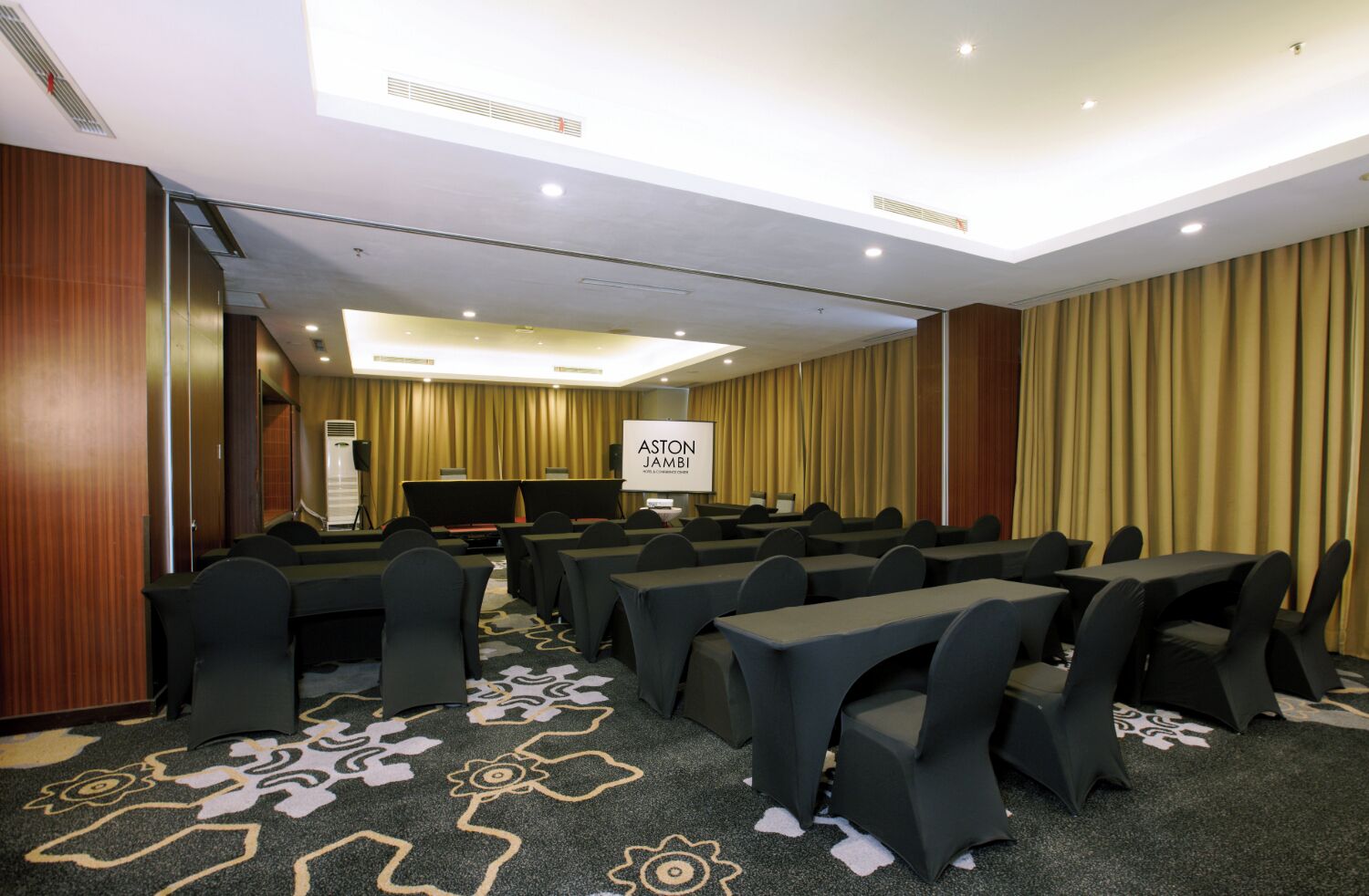 Un salón de conferencias con filas de mesas y sillas, una pantalla de proyector y una alfombra con estampado floral.