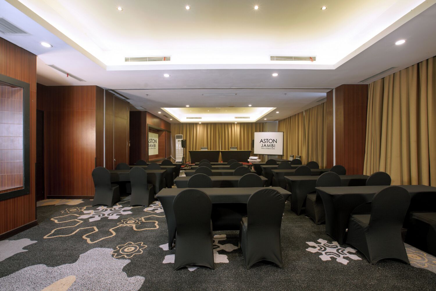 Une salle de conférence équipée de tables et de chaises, d’un écran de projection et d’un sol recouvert de moquette.