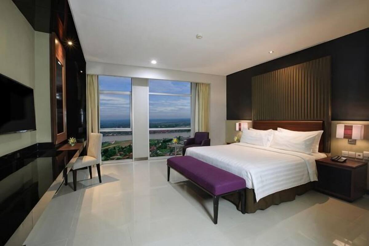 A hotel room with a large bed, a desk, a chair, a TV, and a balcony with a view.