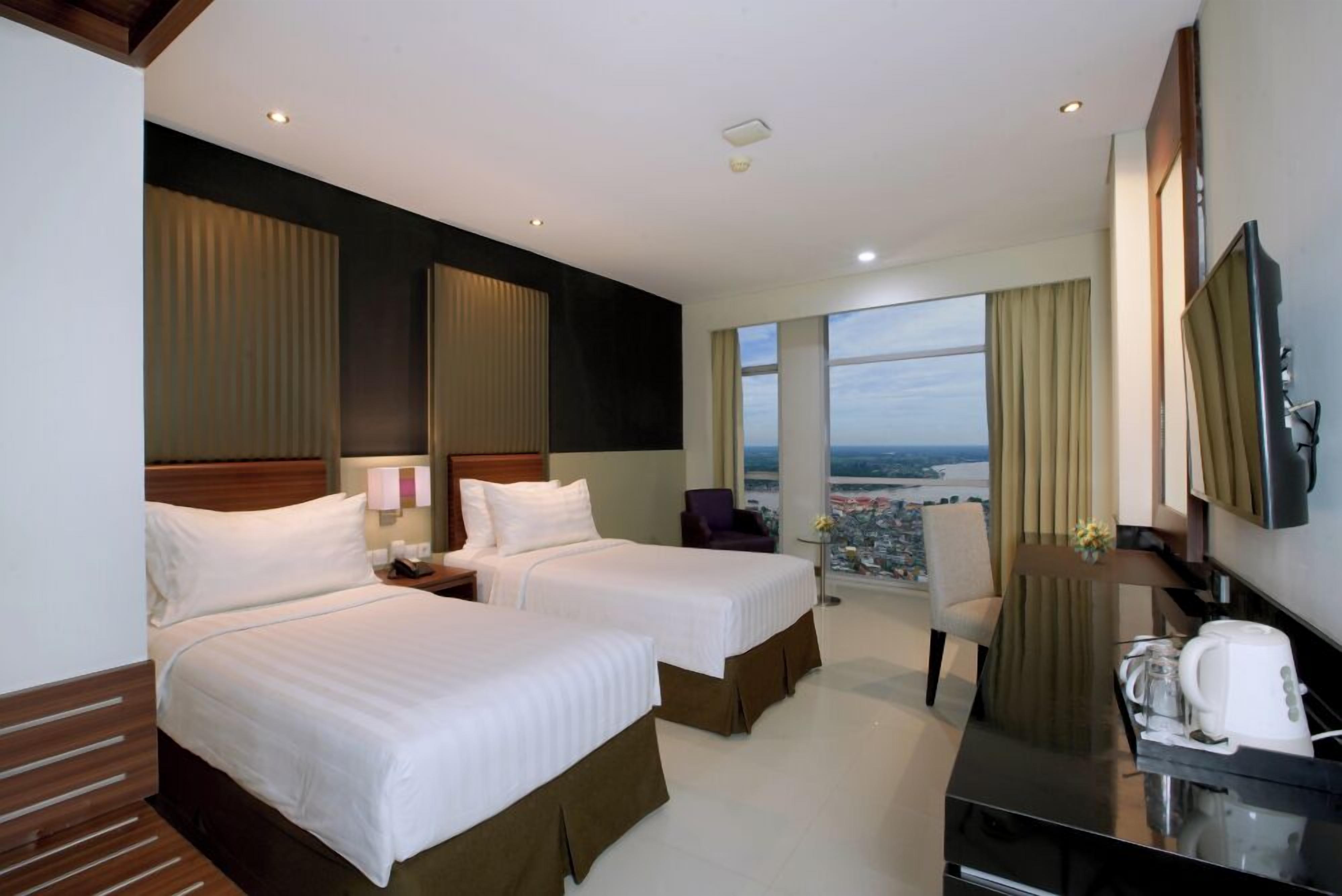 A hotel room with two beds, a desk, a chair, a TV, and a view of the city.