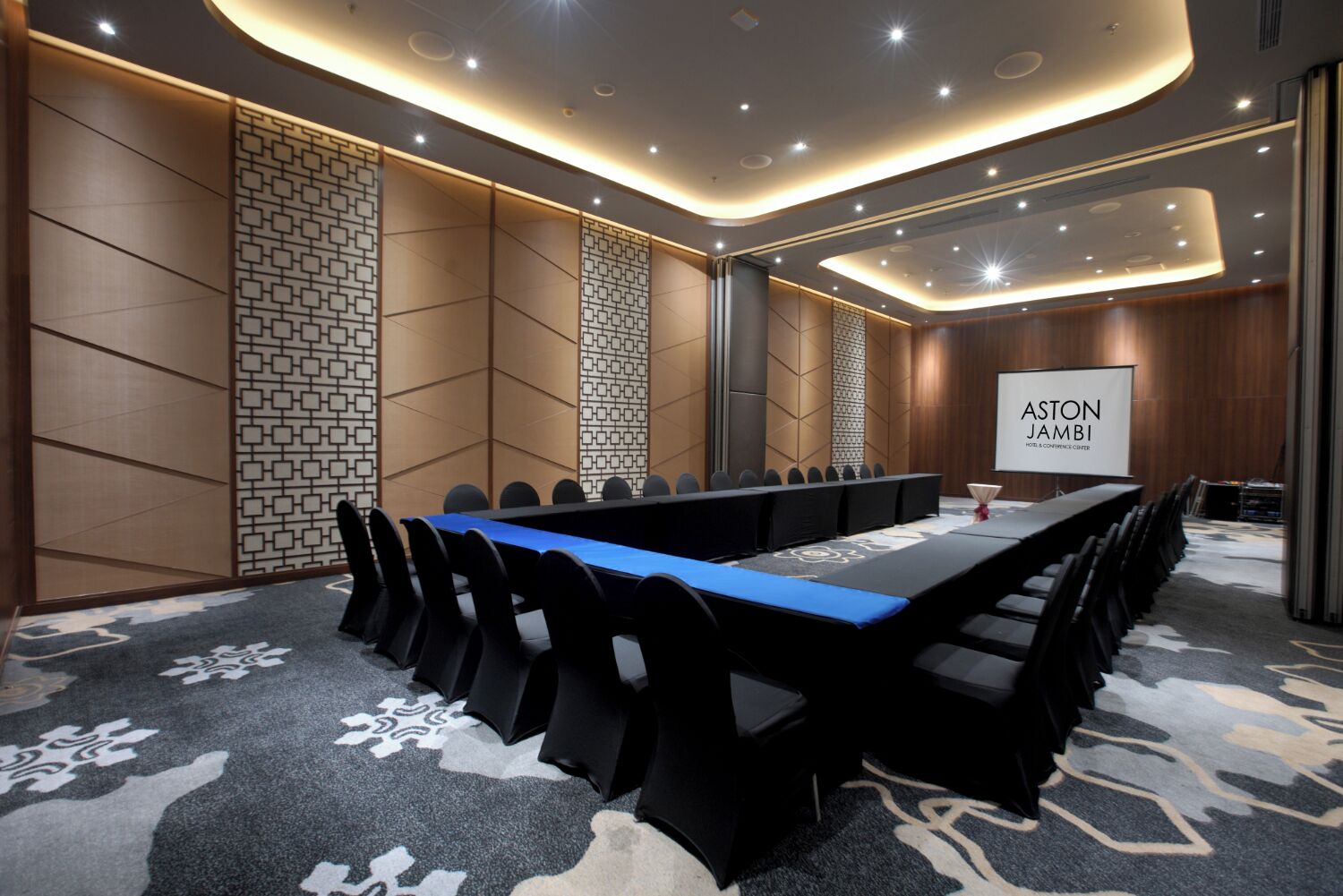 Une salle de conférence avec une longue table, des chaises noires et un écran affichant le texte « ASTON JAMBI HOTEL & CONCIERGE CENTER ».