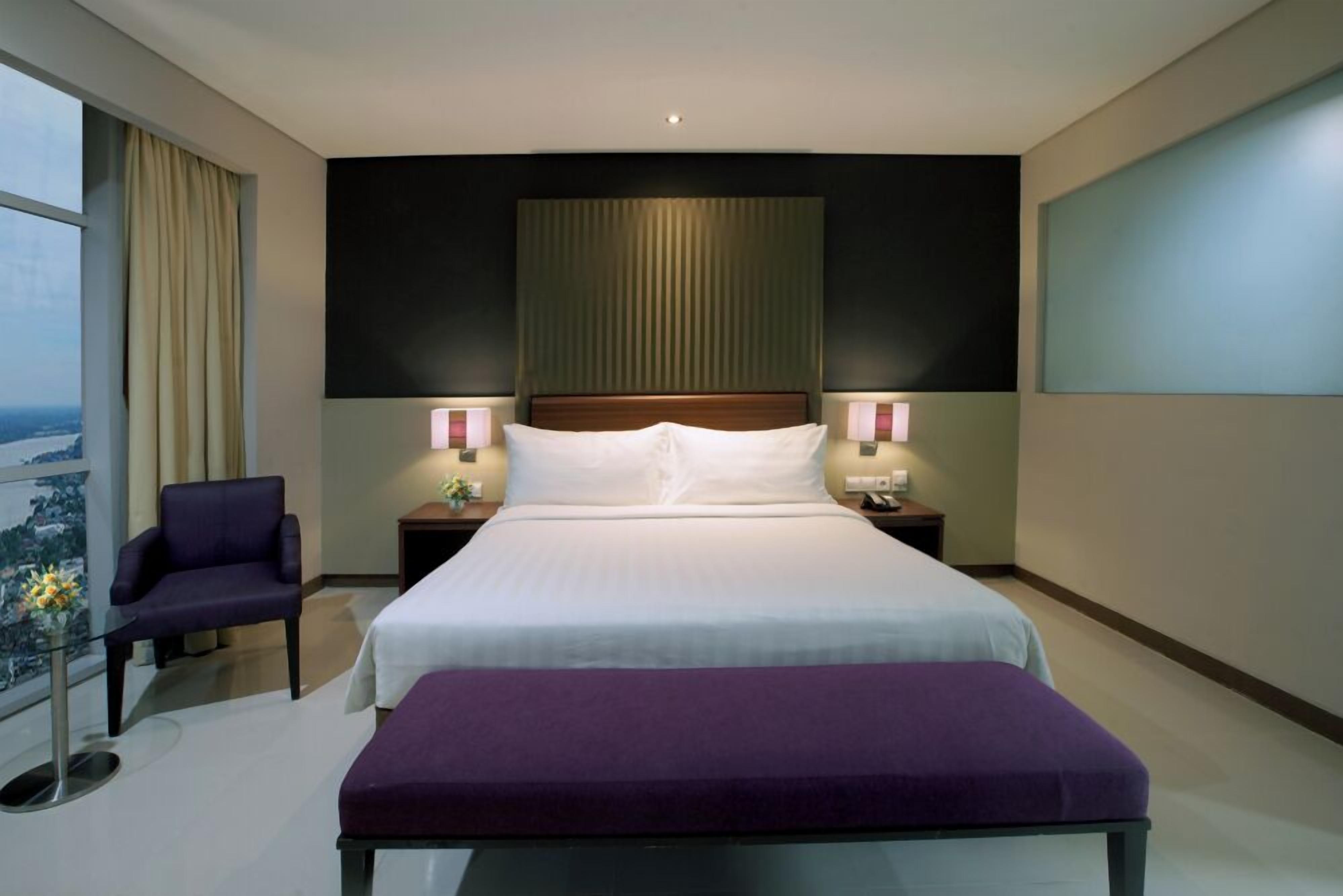 Une chambre d’hôtel avec un grand lit, deux tables de chevet avec des lampes, un fauteuil violet et une vue sur la ville par une grande fenêtre.