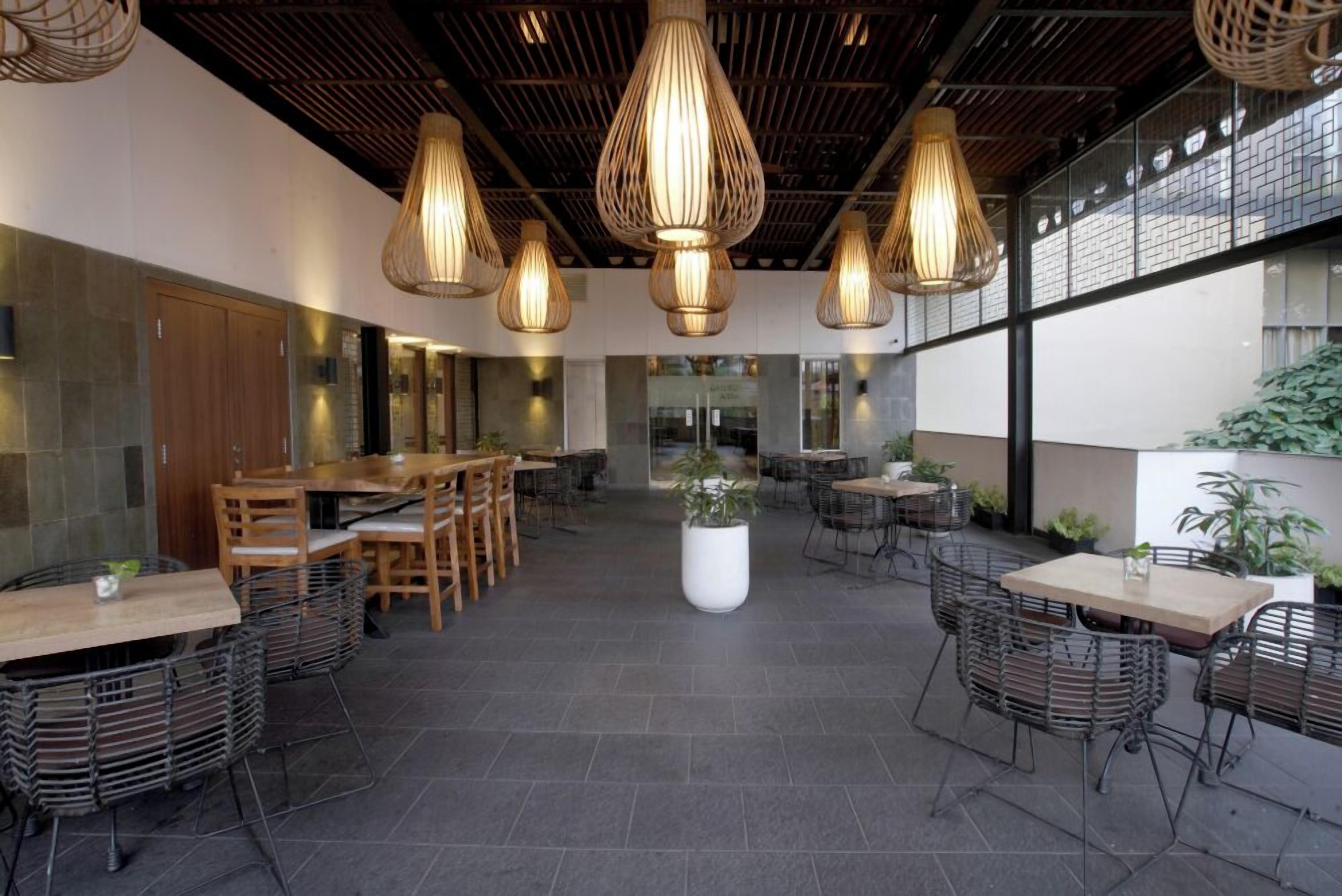 Un comedor interior con varias mesas y sillas, lámparas colgantes y una planta en maceta en el centro.
