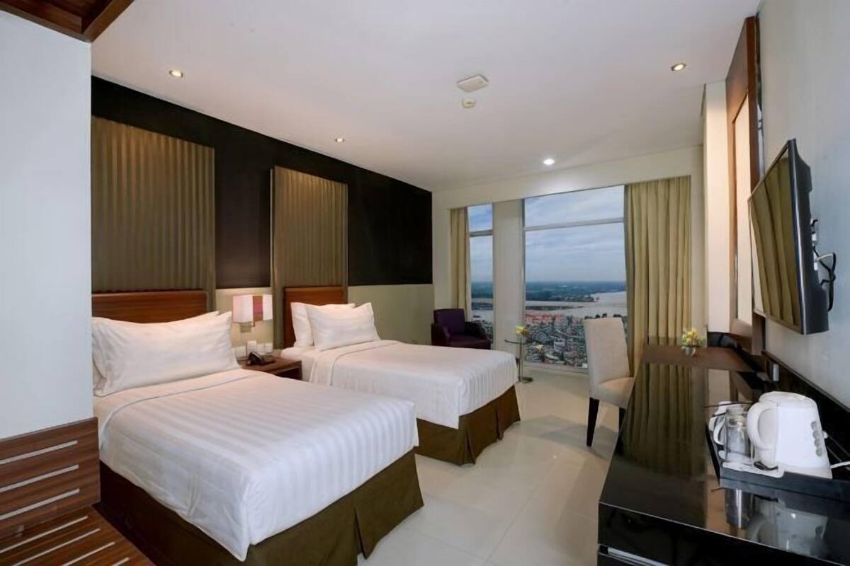A hotel room with two beds, a desk, a chair, a TV, and a large window with a city view.
