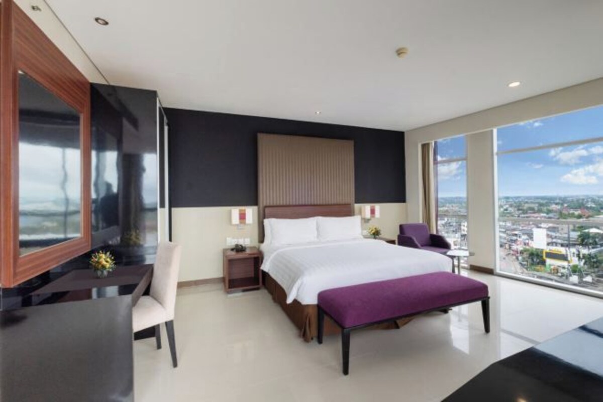 A modern hotel room with a large bed, a desk, and a chair. There is a window with a city view.