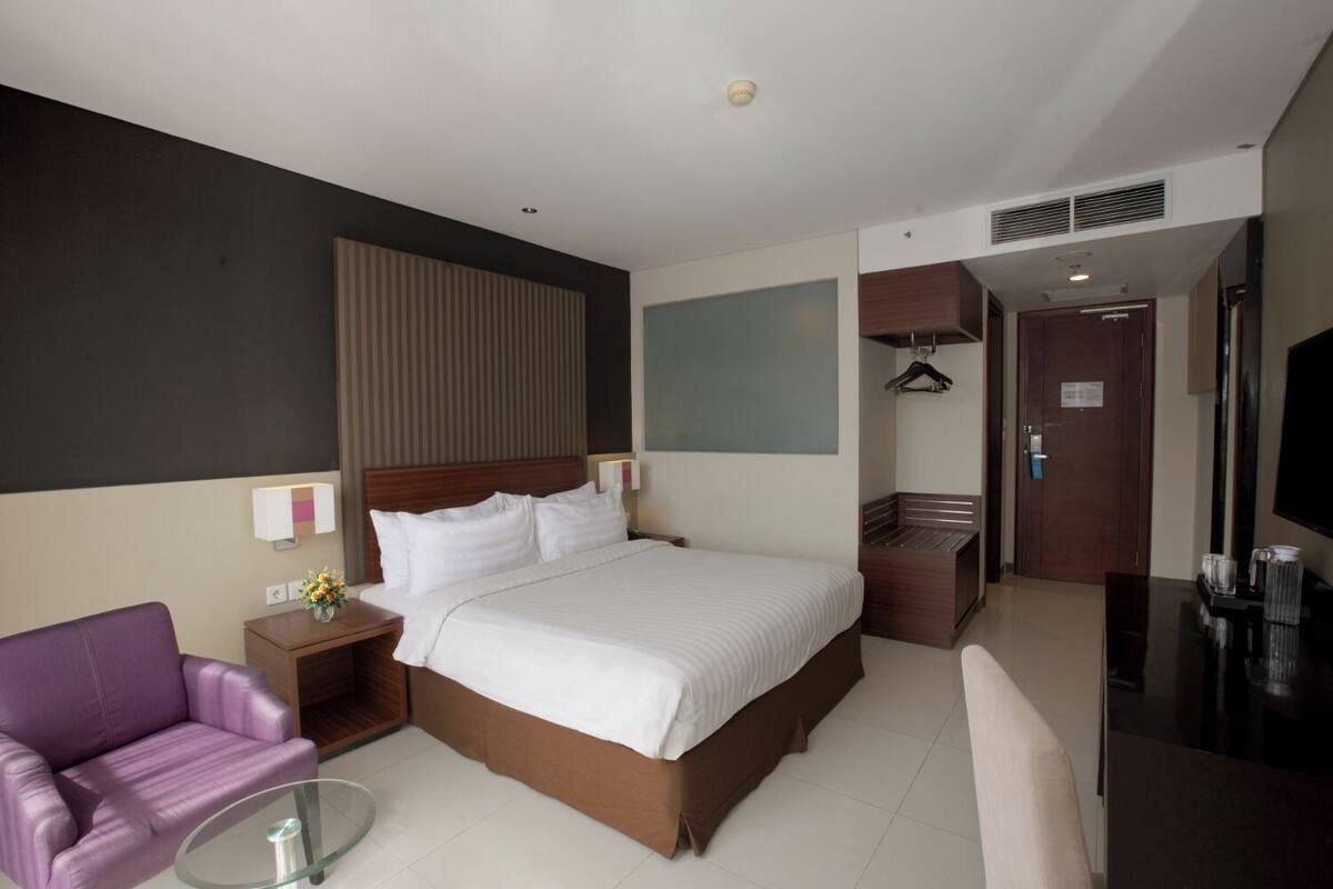 A modern hotel room with a large bed, a seating area with a purple chair, a small table, a TV, and a coat rack.