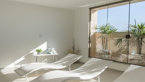Een zonnige kamer met drie witte fauteuils, een klein tafeltje met handdoeken en een plant, en een groot raam met uitzicht op een balkon met beplanting.