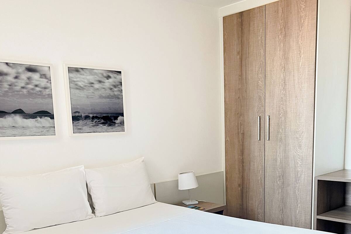 Una cama bien tendida con ropa blanca, un armario de madera con dos puertas y dos cuadros en la pared.