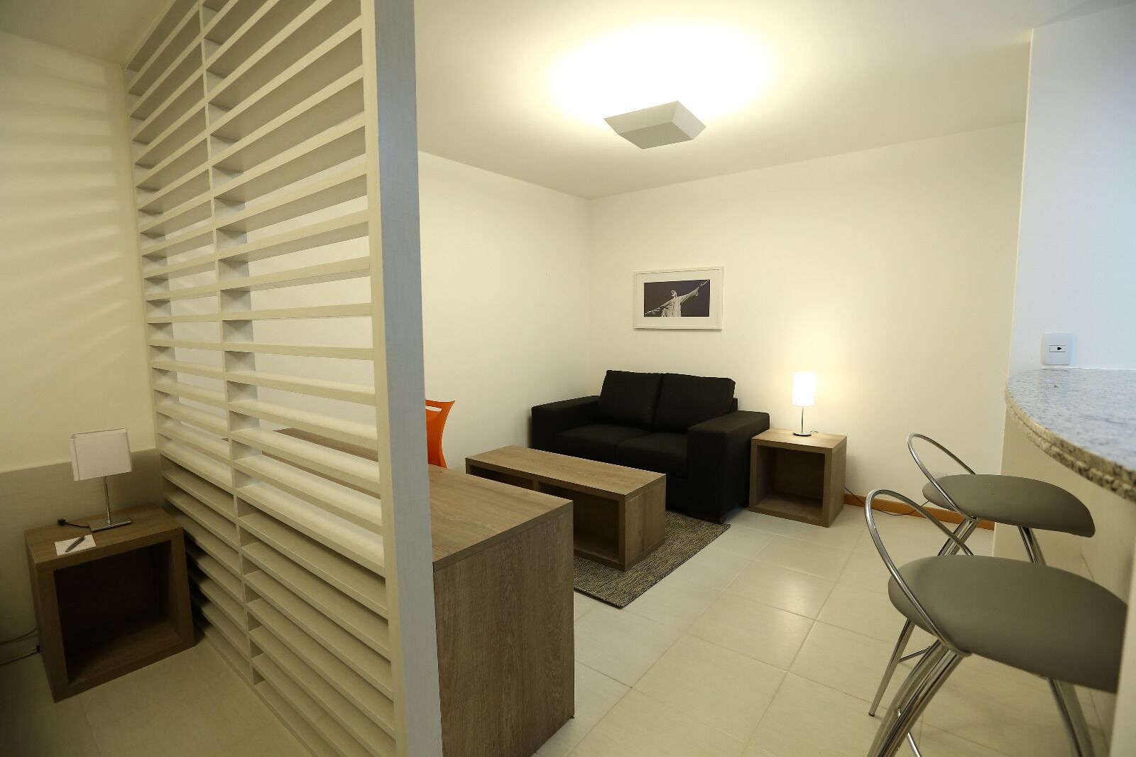 Una sala de estar moderna con un sofá negro, una mesa de centro de madera y un pequeño comedor con dos taburetes de bar.