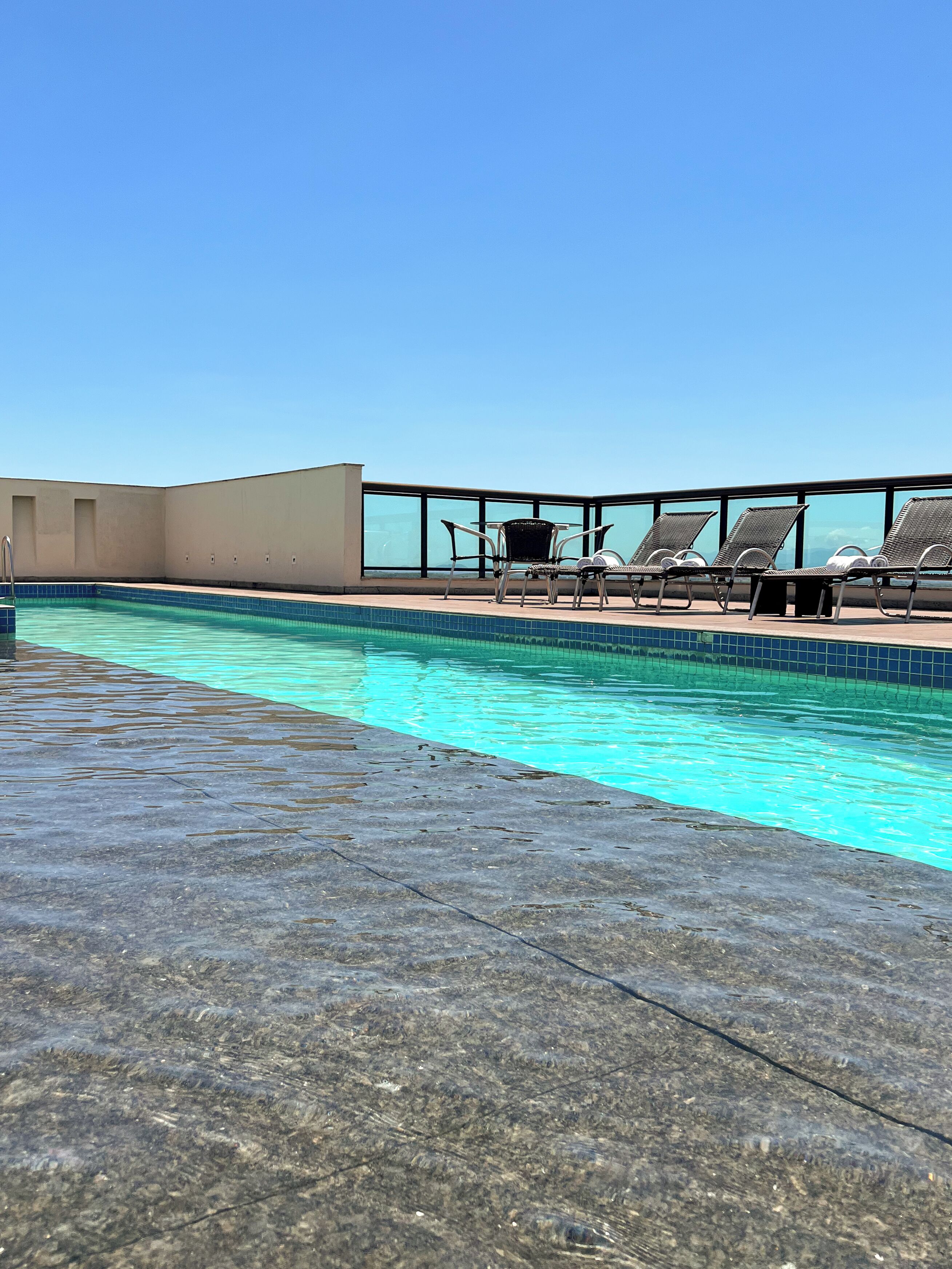 A rooftop pool area with clear blue water, a glass balustrade, and several lounge chairs.