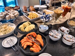 A buffet table with various food items including sliced fruits, pastries, and packaged snacks. - Promenade Prime Itaboraí (Itaborai)