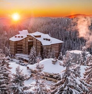 A modern building with a flat roof and multiple windows, surrounded by snow-covered trees and a mountainous backdrop. - Hotel Orlovetz (Chepelare)