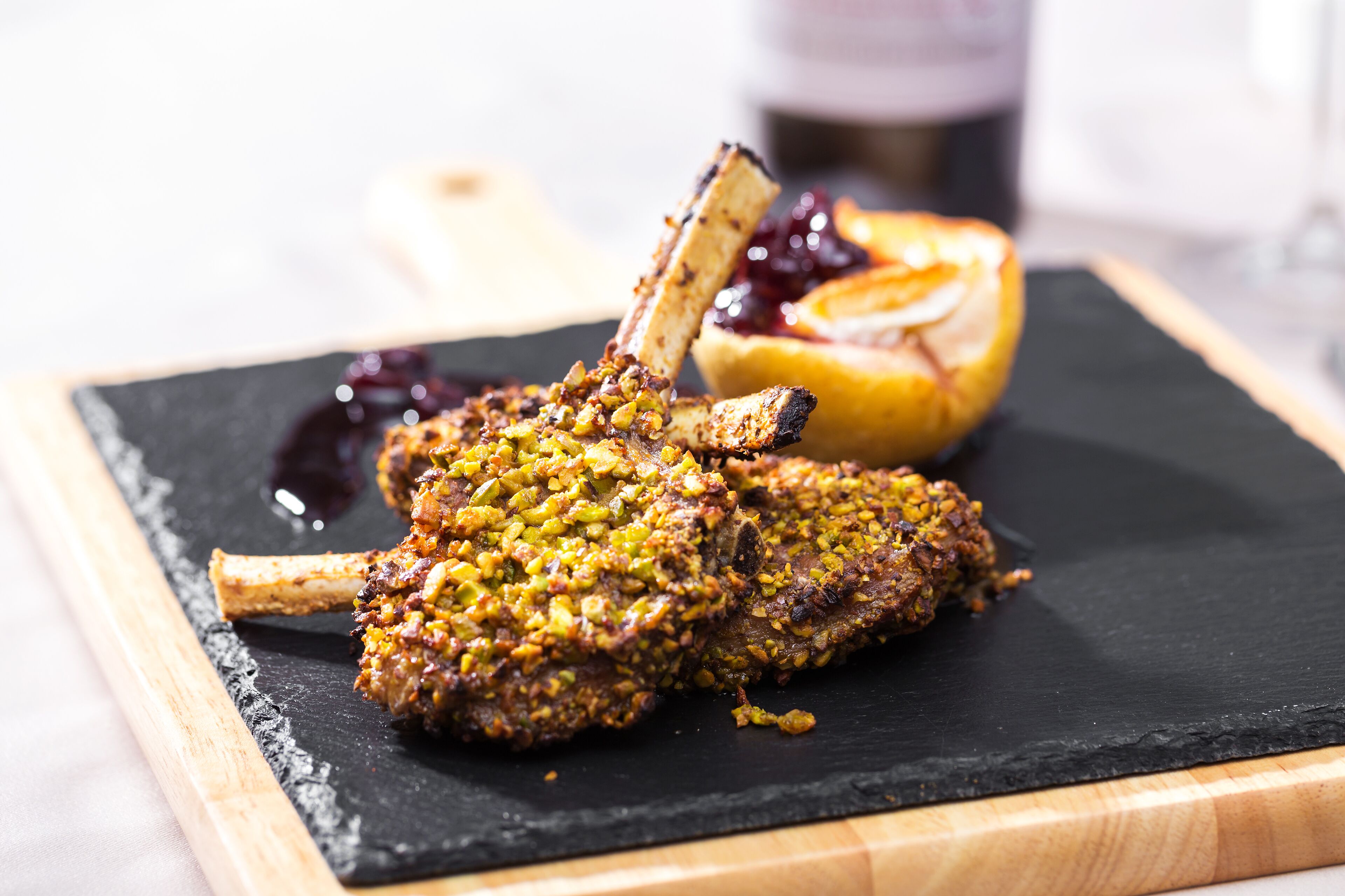 A lamb shank with a crust of herbs and nuts, served on a black slate plate with a side of fruit.