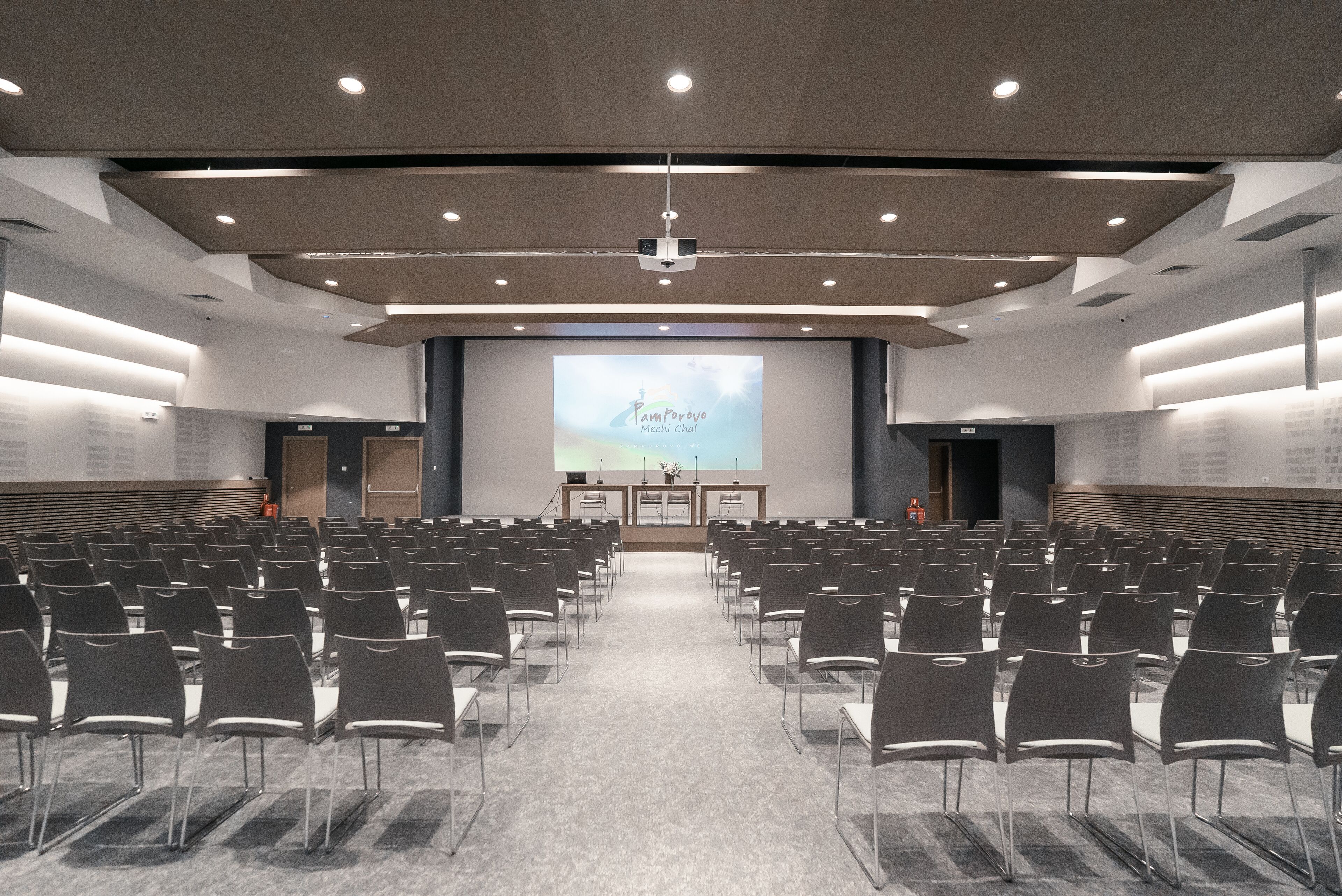 Un salón de conferencias con filas de sillas, un atril y una pantalla grande que muestra una presentación.