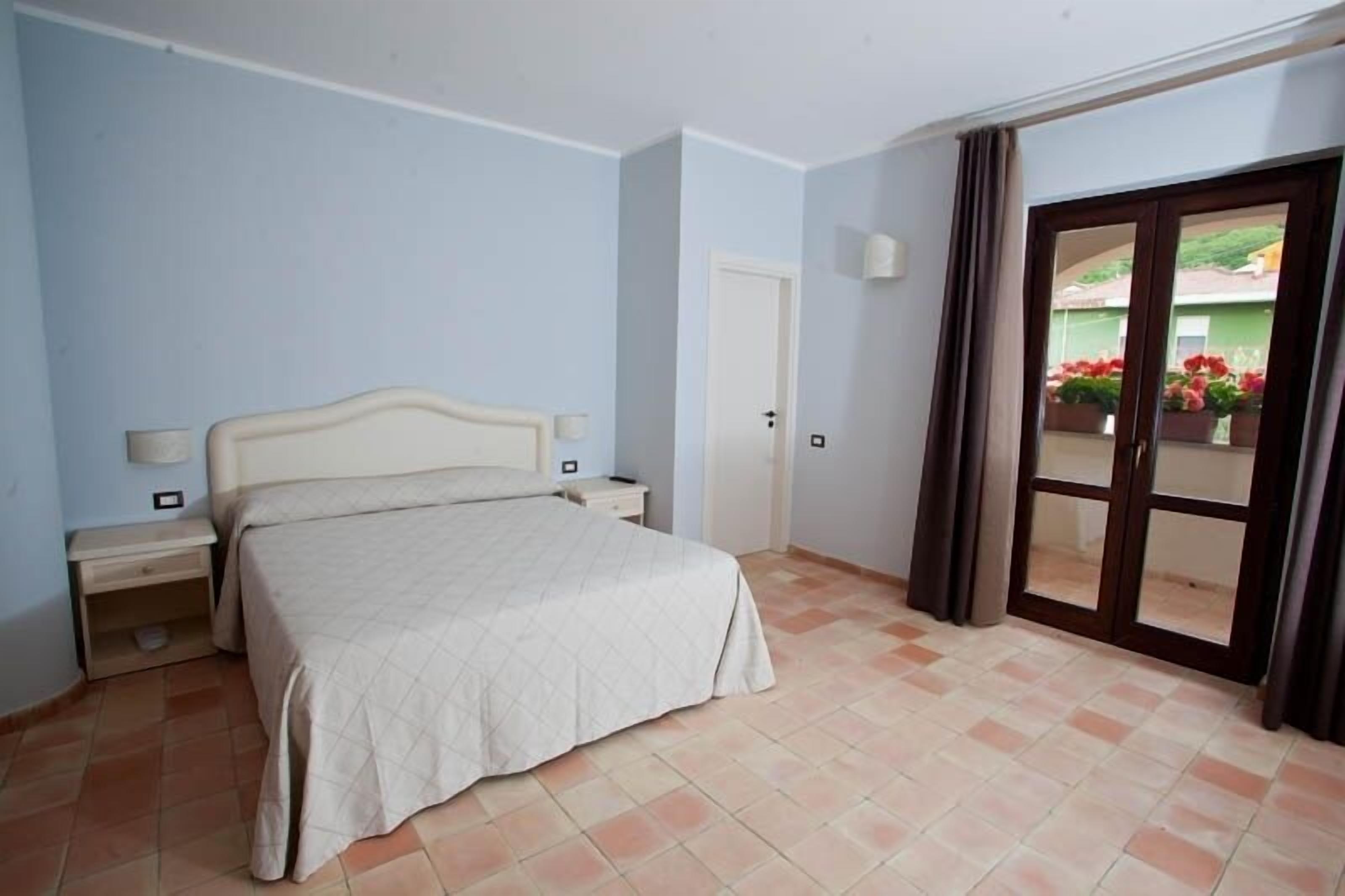 Quarto com cama, mesa de cabeceira e porta que dá para uma varanda com plantas.