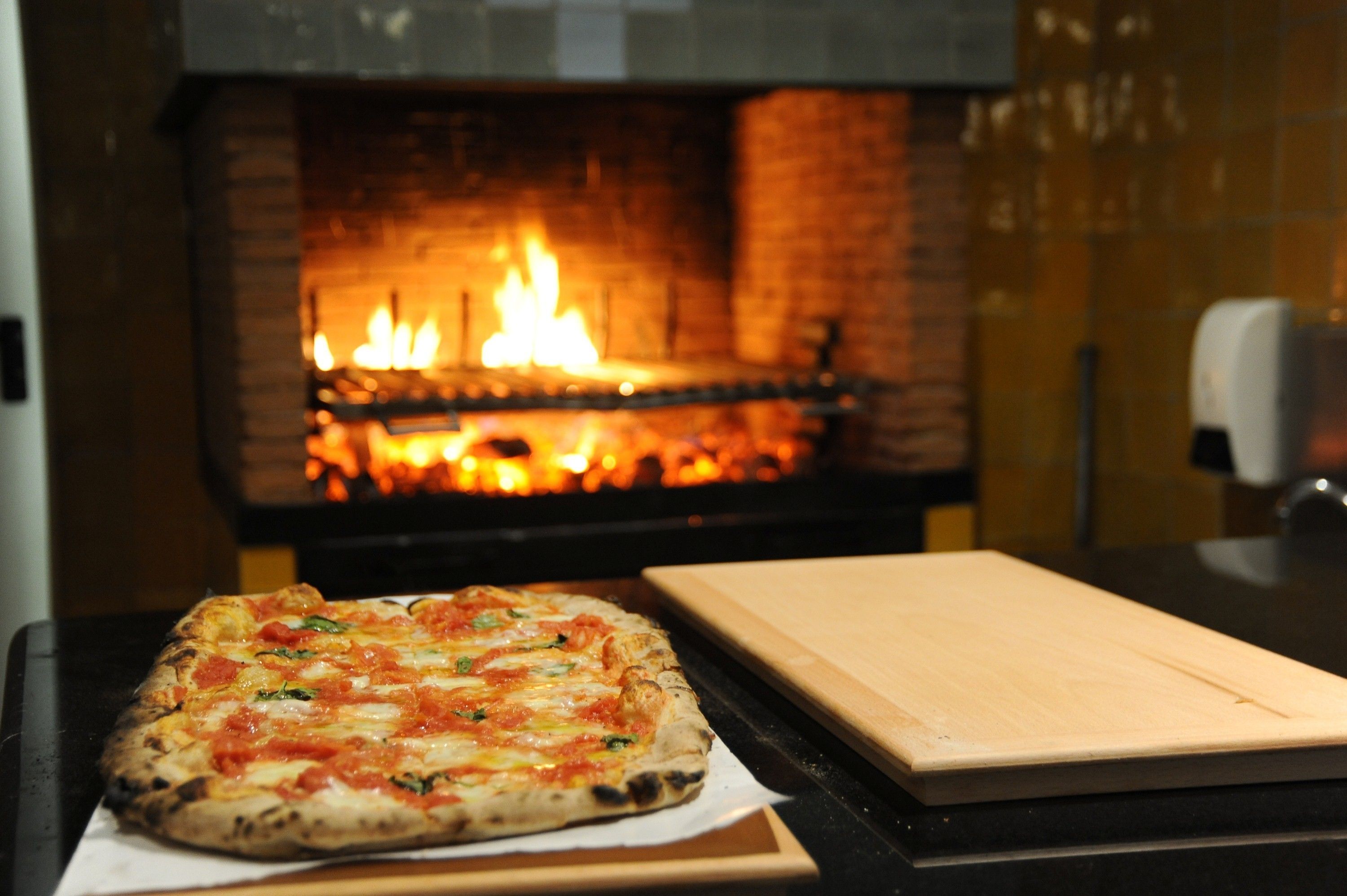 Una pizza sobre una tabla de madera frente a una chimenea de ladrillo con fuego.
