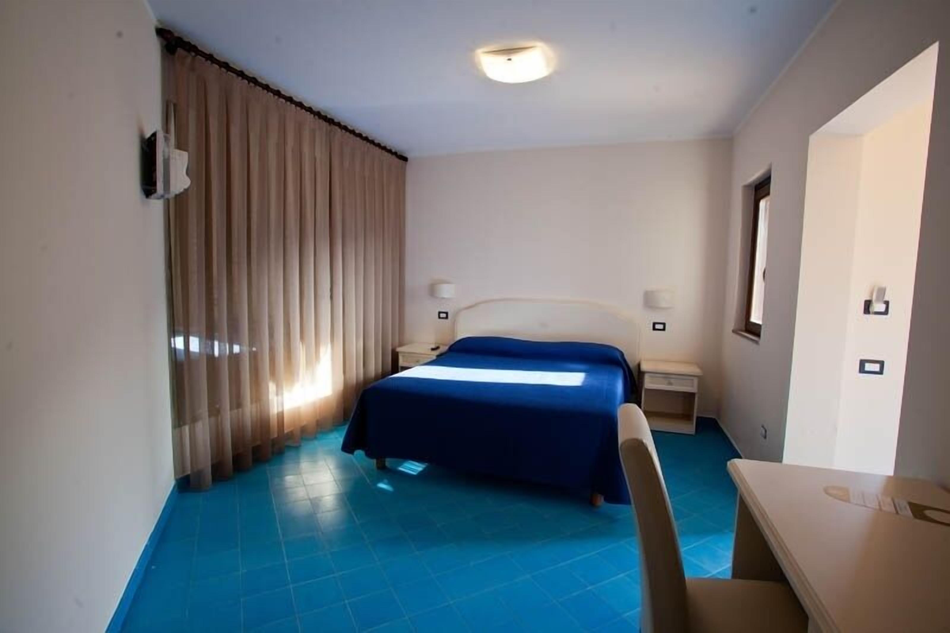 Una habitación de hotel con cama, escritorio, silla, mesita de noche y ventana.