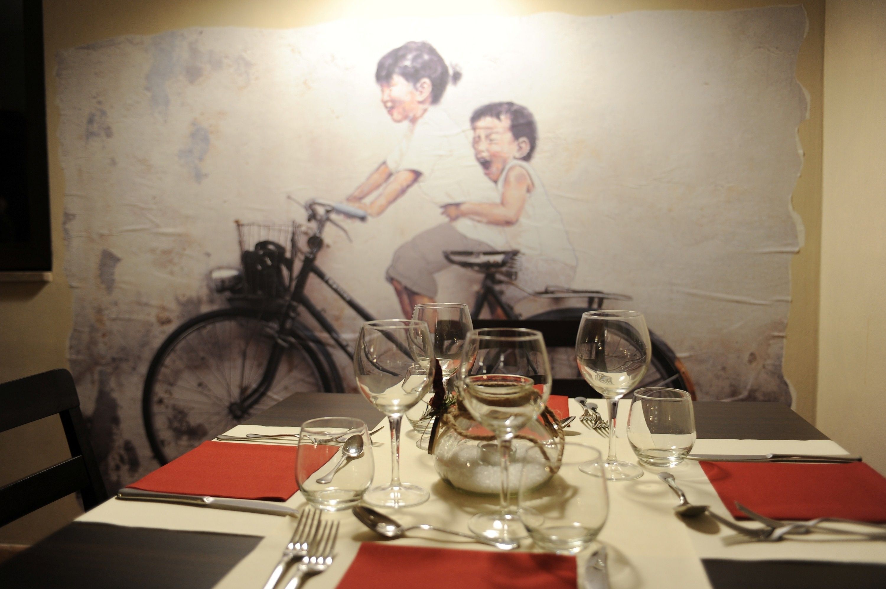 Une table à manger dressée avec des verres à vin, une assiette et des couverts, et une peinture représentant un vélo d’époque en arrière-plan.