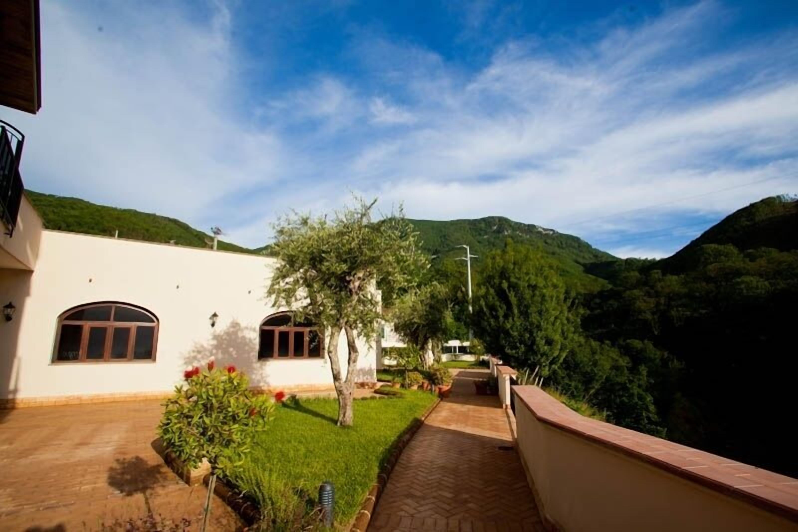 A pathway leading to a building with a white exterior and arched windows, surrounded by greenery and hills.