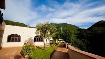 A pathway leading to a building with a white exterior and arched windows, surrounded by greenery and hills.