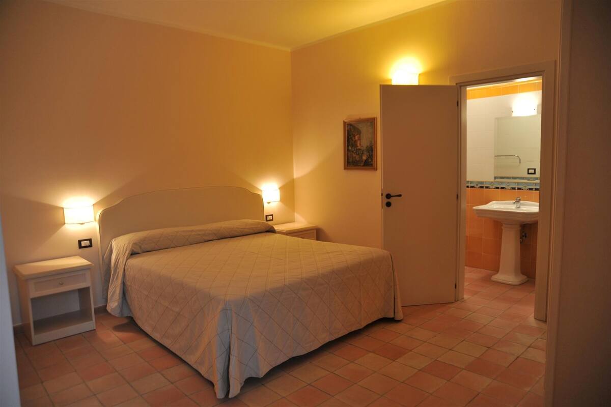 Une chambre à coucher comprenant un lit, des tables de chevet et une porte donnant sur une salle de bain équipée d’un lavabo et d’un miroir.
