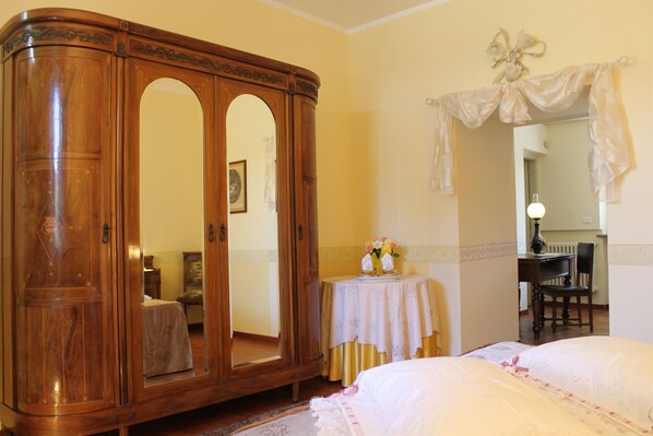 A room with a large wooden wardrobe, a bed with white bedding, a small table with a vase of flowers, and a chair.