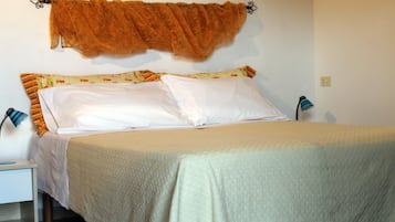 Un letto rifatto con precisione, con copripiumino beige e due cuscini bianchi, un comodino con una lampada e una tenda fissata a parete.