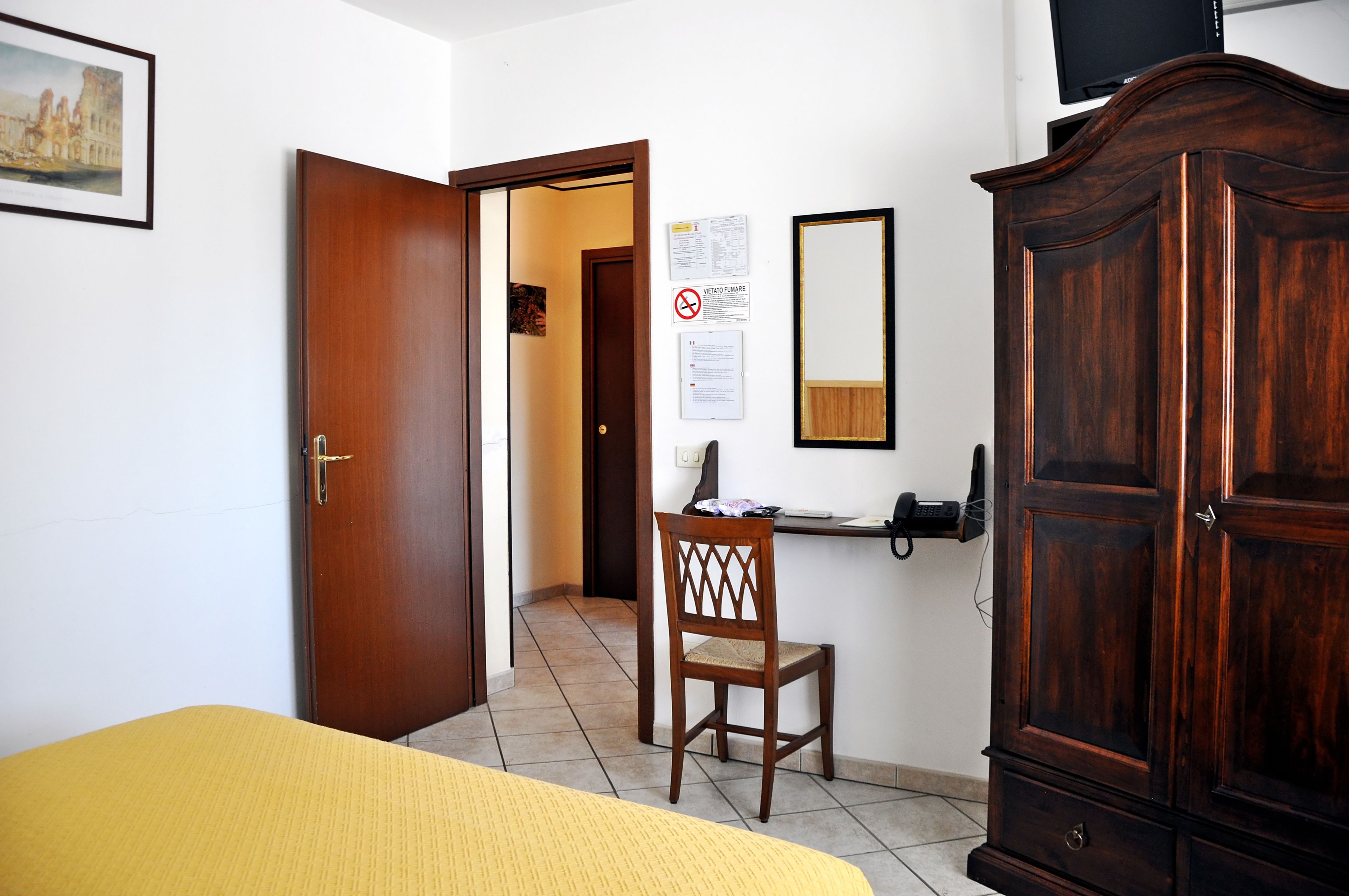 A room with a bed, a wooden wardrobe, a chair, a desk with a telephone, and a door leading to another room.
