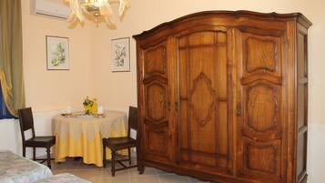 Ein Zimmer mit einem großen Holzschrank, einem Esstisch mit gelber Tischdecke, zwei Stühlen und einem Bett mit blumigem Bettzeug.