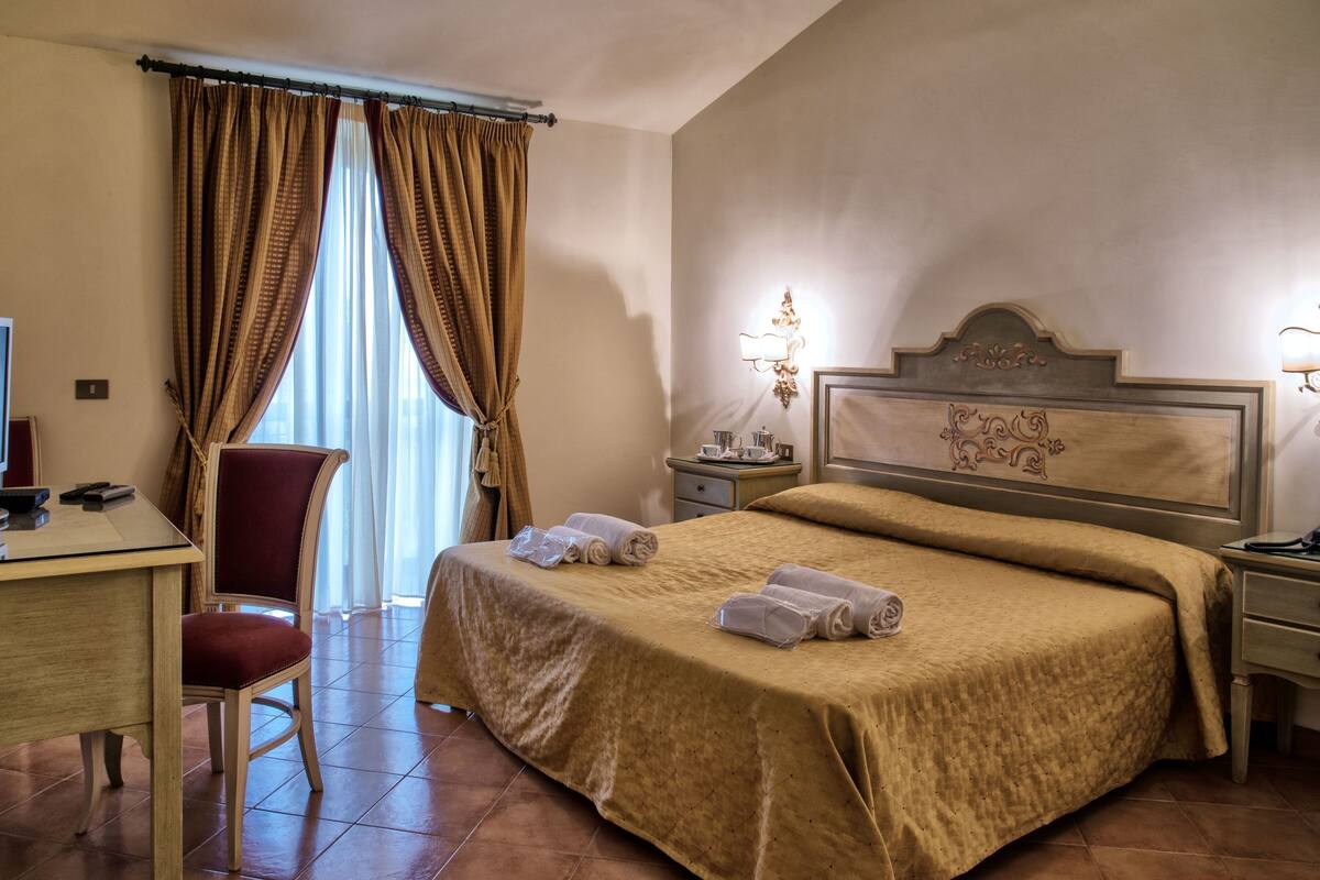 A hotel room with a bed, a desk with a computer, a chair, and a nightstand with a lamp.