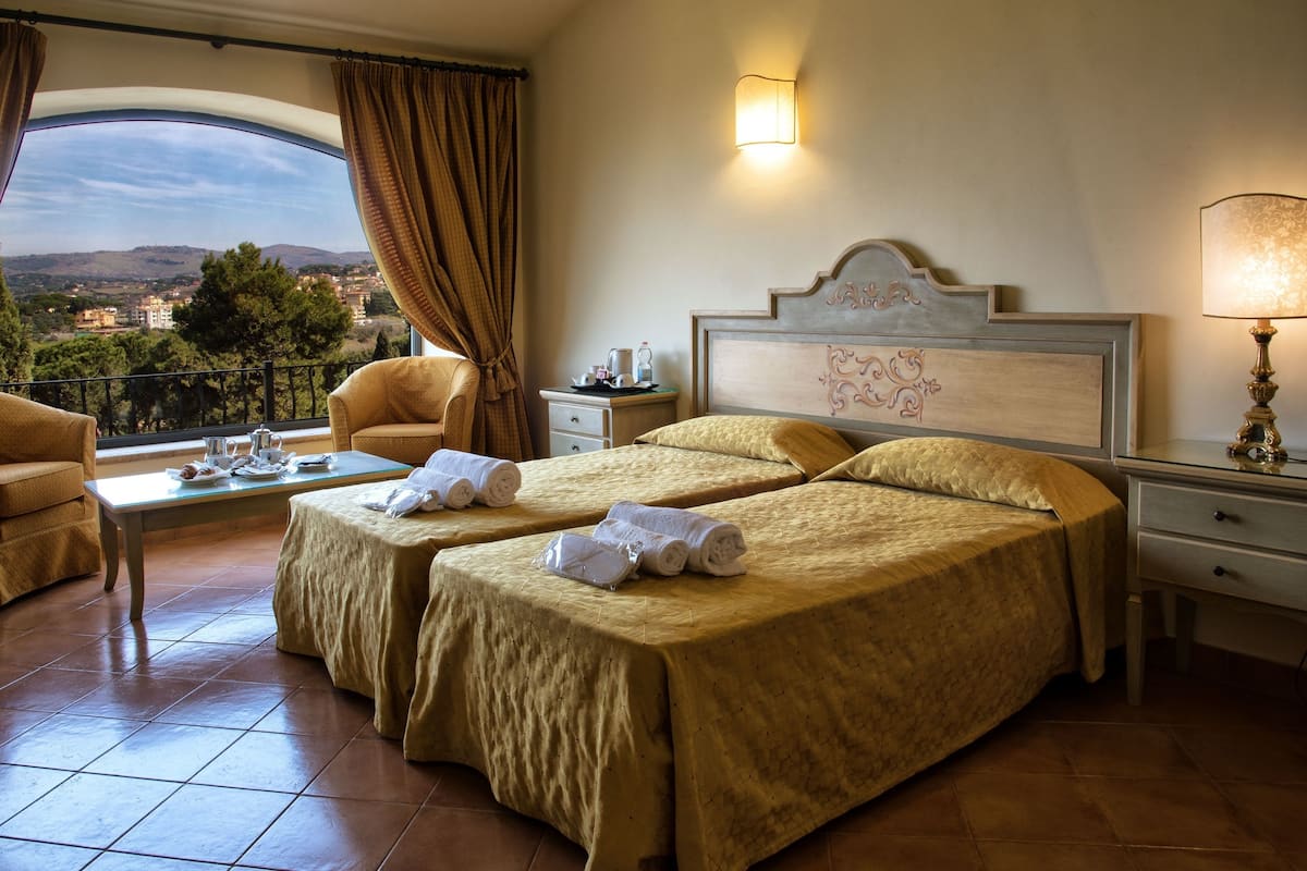A hotel room with two beds, a chair, a table with a view, and a nightstand with a lamp.