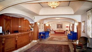 A lobby with a reception desk, blue sofas, a red sofa, and a chandelier. - Grand Hotel Helio Cabala (Marino)