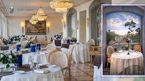 A Grand Hotel Helio Cala dining area with multiple round tables, chandeliers, and a scenic view through an arched window. - Grand Hotel Helio Cabala (Marino)