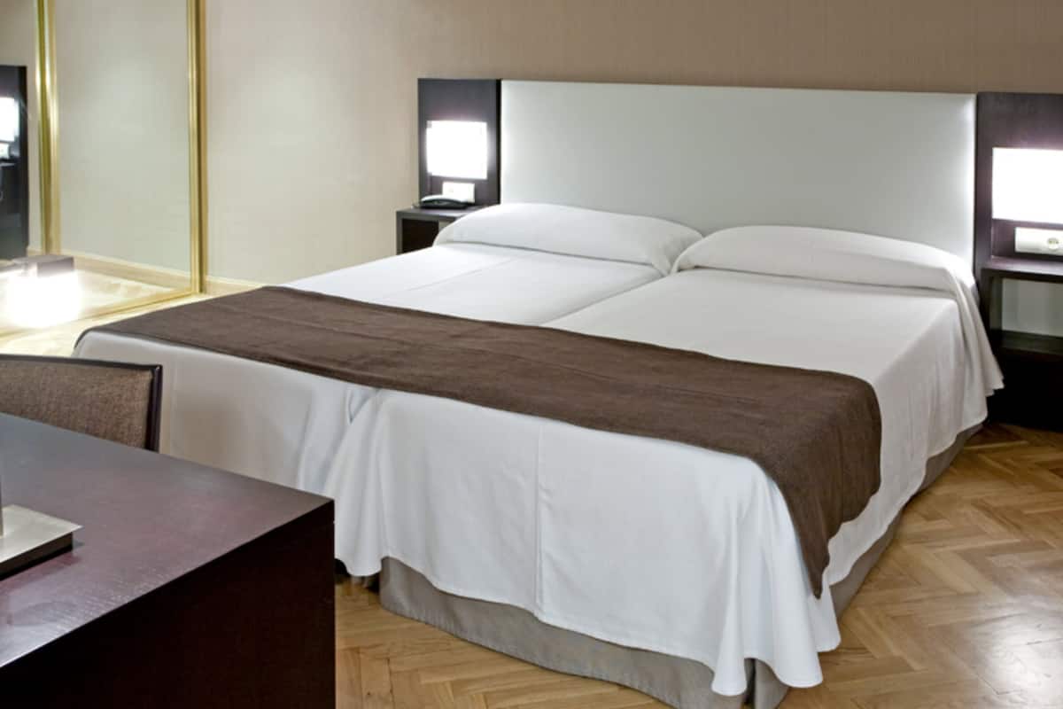 A double bed with a white and brown bedspread, two bedside tables with lamps, a wooden floor, and a large mirror on the left wall.