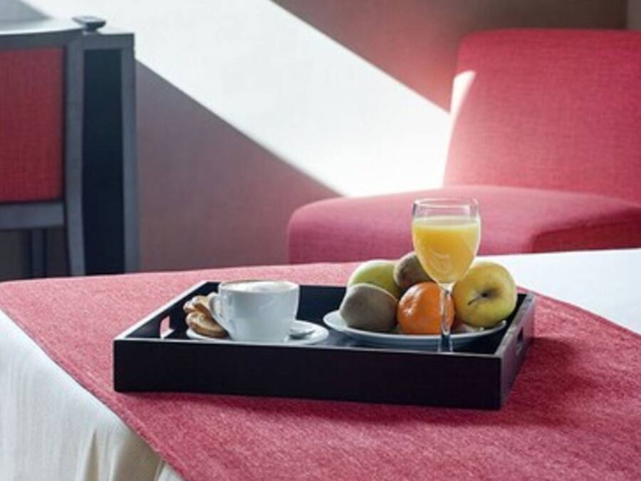 A breakfast tray with a cup, pastries, and fruit on a bed with a red chair and a nightstand in the background.
