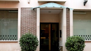 Entrada de hotel con puerta de vidrio, un pequeño toldo y dos plantas en macetas.