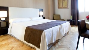 Habitación de hotel con una cama grande, dos mesitas de noche con lámparas, una silla, una pequeña mesa y una ventana con cortinas.